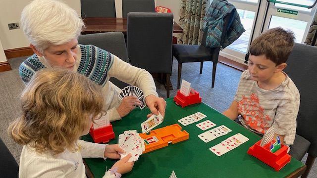 Rebecca Moore playing bridge with two children, a boy and a girl. They are sitting around a table with a green top on it, and one child is holding a number of playing cards while the other child is looking at cards in front of him. Rebecca has short white hair and is wearing a patterned jumper. The girl has wavy shoulder-length hair and the boy has short dark hair and is wearing a T-shirt. There are chairs and a table behind them and a door