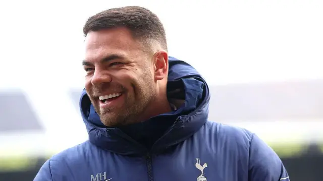 Martin Ho laughing as he wears a hooded blue coat with Tottenham emblem and letters MH above Nike logo