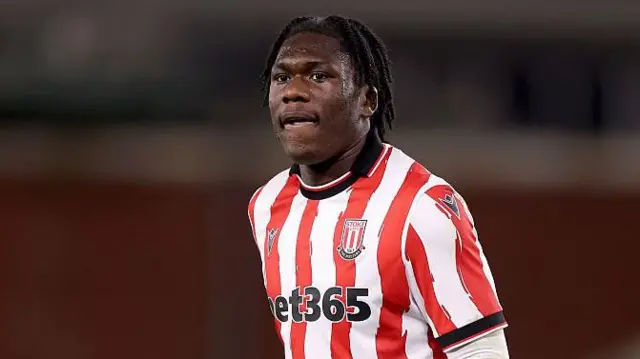 Sydney Agina standing on the pitch with his arms by his side, wearing a Stoke City red and white striped home shirt