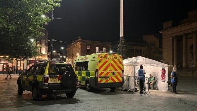 Photo shows a police car and an ambulance parked beside a white tent in Barker's Pool, a public square in Sheffield city centre. It is night time and in the distance there are two people stood outside the tent and once person sitting inside waiting to help people.