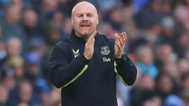 Sean Dyche, wearing a dark-coloured Everton top while in charge of the Blues, applauding towards his players in encouragement on the touchline during a game, with the crowd out of focus in the background.