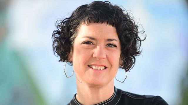 Laura Murphy, a young woman with black curly hair and octagonal silver earrings, smiles at the camera against a neutral, light blue background