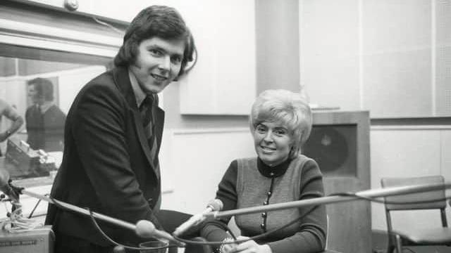 Black and white photo of George Hamilton sitting on a desk with microphones on it wearing a suit and tie. Gloria Hunniford is sitting next to him at the desk.