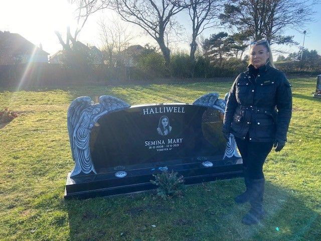 Rachel Halliwell stands in front of her daughter's grave. The grave is made from black stone and there is a photo of Semina in the centre. There are angels to each side of the headstone.