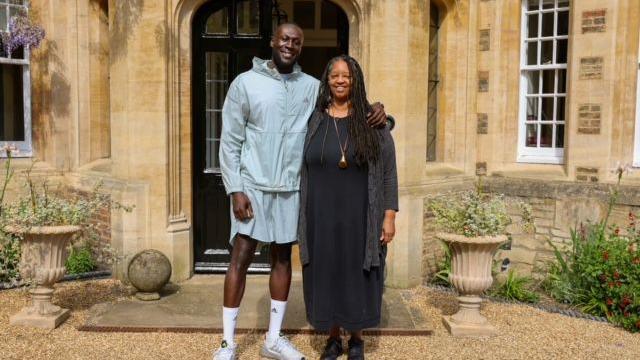 Stormzy is standing alongside Sonita Alleyne OBE outside a classical building entrance, framed by arched black doors and decorative planters. Dressed casually in a hooded jacket, shorts, and white sneakers, Stormzy poses next to Sonita, who wears a long dark dress. The pair smile for the camera, marking Stormzy’s election as an Honorary Fellow of Jesus College, Cambridge
