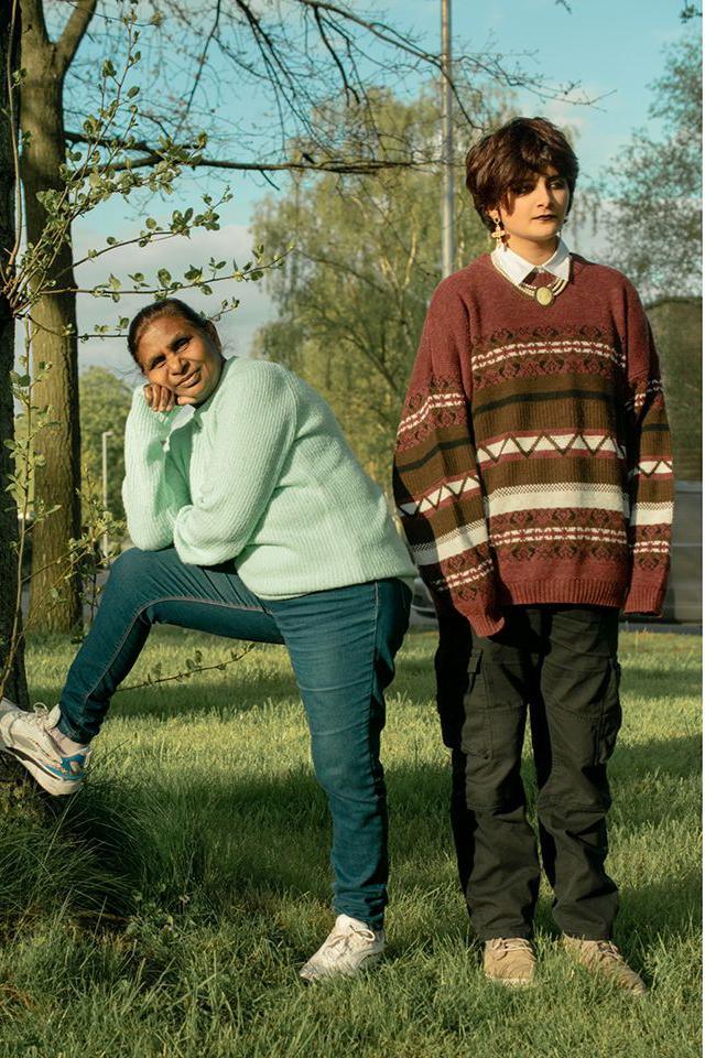 An older woman wearing jeans and jumper with one foot on a tree stump. Next to her a taller woman dressed in trousers and a jumper stands looking ahead. 