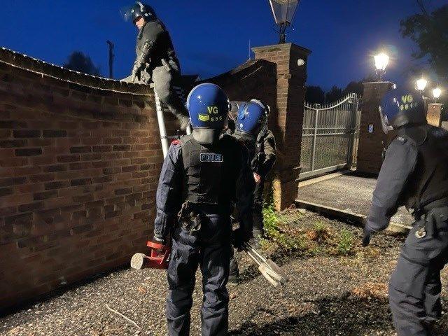 Officer in protective clothing and helmet straddles a wall after climbing a silver ladder. Behind them are three other officers, with their back to the camera, looking on