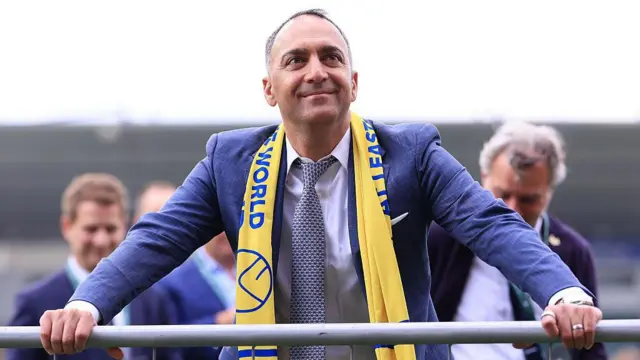 A smiling Paraag Marathe holds onto a barrier at Plymouth Argyle's Home Park after Leeds win the Championship. He has a yellow scarf round his neck