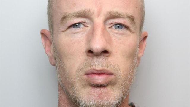 A mugshot of a middle-aged man with a a short grey beard and grey/blond closely cropped hair.