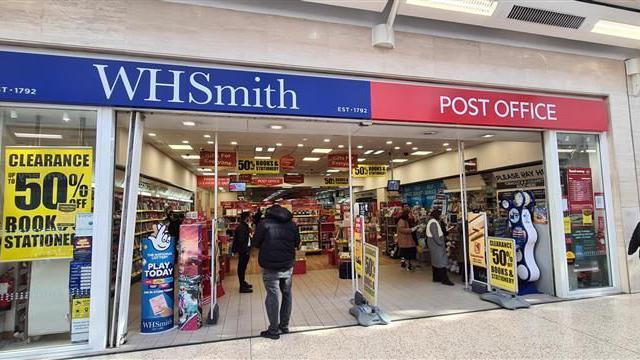 A view of the WHSmith store in Luton Point. It is open with customers inside. There is red post office signage above the entrance.