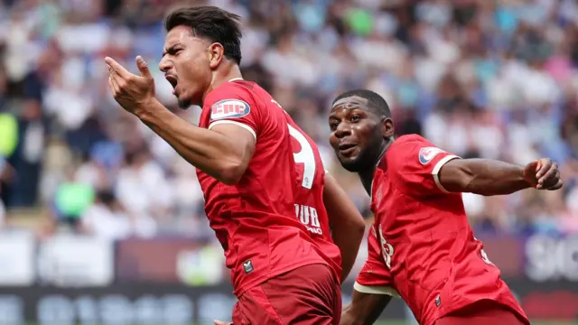 Jusef Erabi, wearing Preston's red away shirt, celebrates scoring against Bolton by raising his left arm in the air. Odel Offiah can be seen next to him.