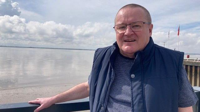 A bald man, with glasses is standing in front of a railing by the river Humber. He is smiling at the camera and wearing a navy blue t shirt and navy blue body warmer.