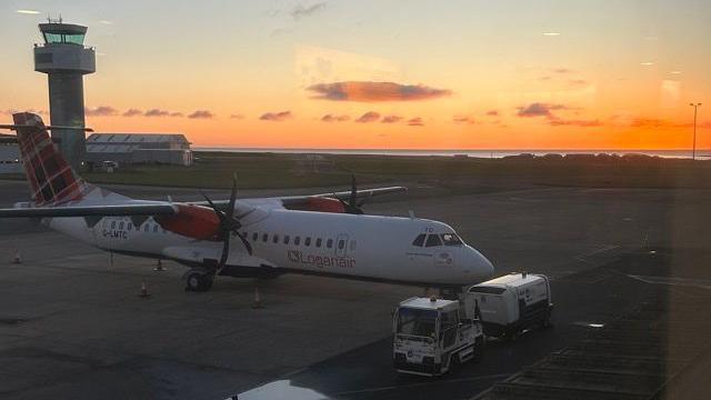 A Loganair plane on the tarmac at Ronaldsway at sunrise. There is a small vehicle for transporting luggage parked in front of it .