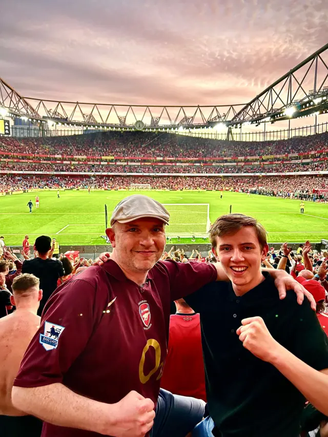 Michael and his Dad standing at the Emirates Stadium watching Arsenal 