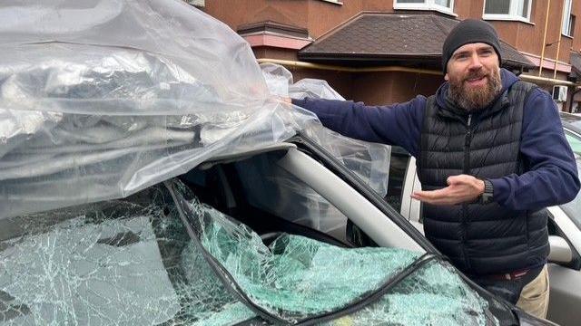 Oleksandr Vilko, who has a beard and is wearing a black vest and beanie, points to his car, which has a shattered windshield.