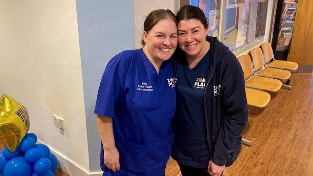 Katy Jones and Laura Knight, in blue scrubs, smile and pose for the camera with their faces touching 