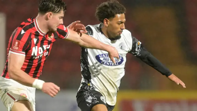 Tyrelle Newton in white Gateshead shirt, with black undershirt sleeves and shorts, battling with a Walsall player on the left with red and black stiped shirt