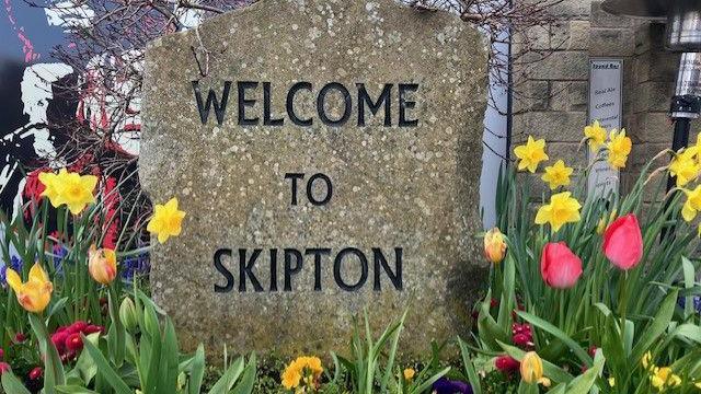 A stone sign saying 'Welcome To Skipton' in black letters surrounded by brightly coloured flowers - red tulips and yellow daffodils.