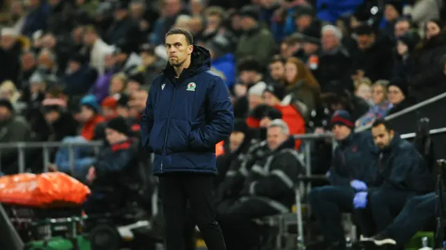 Valerien Ismael watching Blackburn Rovers wearing a navy blue puffer jacket and black trousers with the crowd in the background