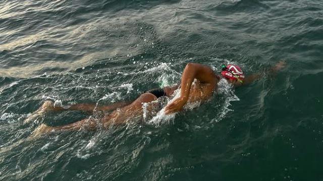 Premal Pattani wearing black Speedo swimming trunks a red neoprene hat and goggles. He is swimming front crawl in the open sea at dusk, and the water is a deep dark green colour.