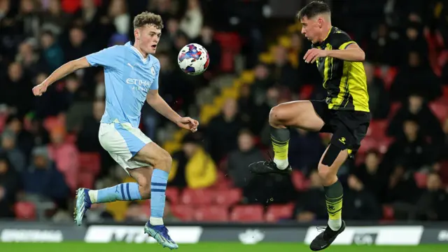 Two footballers jumping for the same ball. One on the left wearing a light blue Manchester City kit and the other wearing a black and yellow Watford kit