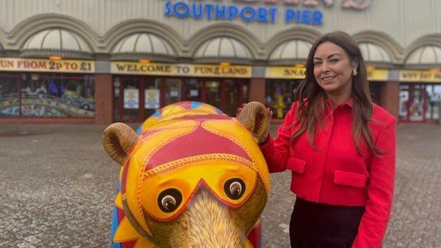 Serena Silcock Prince stands in front of the Southport Pier amusements building. She is standing next to a statue of a bear decorated in bold primary colours. She wears a red jacket and black trousers and has long dark hair which she is wearing down as she holds the bear's ear and smiles at the camera.