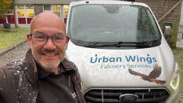 Andy Crowle smiling for the camera in front of the Urban Wings white company van. There is a hawk drawn on the bonnet. It is snowing. There is a building behind him.