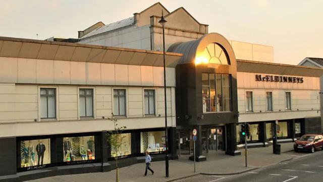 A high street department store photographed in early morning sun. The sun is reflecting off its glass frontage, a single shopper walks by.