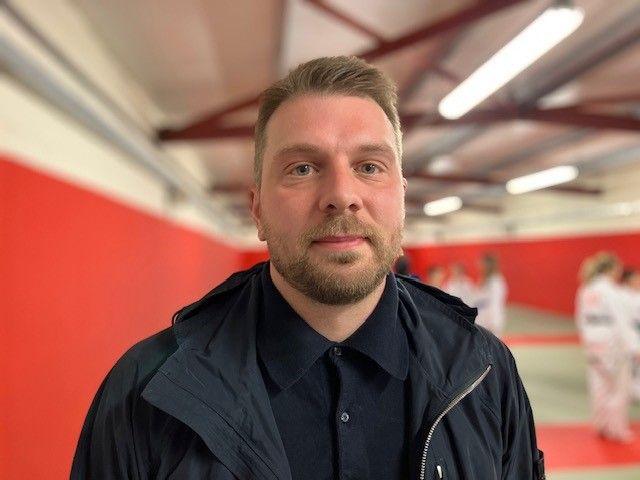 Jamie Stringer has short sandy brown hair and a close cropped beard, he is wearing a dark blue shirt and jacket and stands in front of people in a gym who are wearing martial arts uniforms.