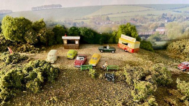 A close up of a model hillside with vintage cars parked along with a small wooden stall and ice cream van. There are shrubs and trees and more rolling hills seen in the background