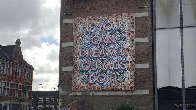 If You Can Dream It You Must Do It artwork in Luton. The artwork is on a brick building, surrounded by other brick buildings. The working is in a red colour, with a background in blue and orange colours