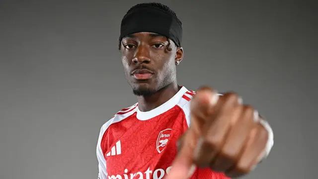 Noni Madueke wears an Arsenal shirt and points at the camera up close