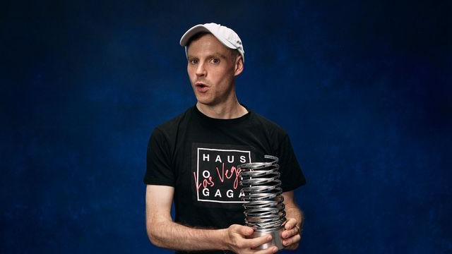 A man with a black T-shirt which teads "Haus of Gaga" holds a metal award like a coiled spring on a metal base. He wears a white baseball cap.