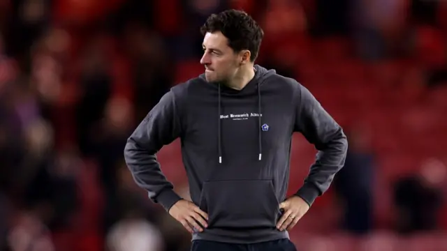 Ryan Mason looks to his right with a forlorn look on his face and his hands on his hips. he wears a grey hoodie.