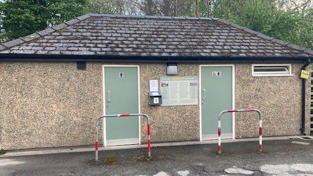 A single storey, grey pebble dash toilet block, with two green doors and a slate tile roof. Behind the building is a row of trees 
