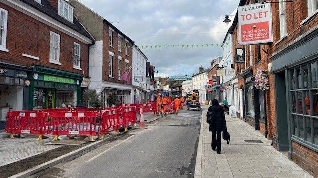 A single road in a high street with shops either side. The road jas red fences up and people in orange hi-viz suits working on it.