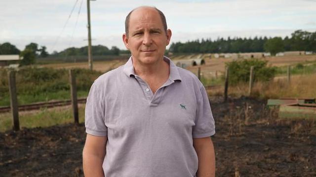 William Edwards is wearing an open-necked blue shirt and standing in front of a wooden fence, fields and farm buildings.