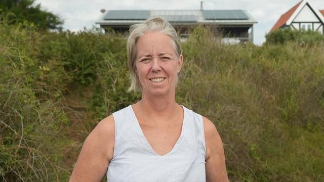 Sophie Marple a woman who is standing in front of a cliff top with shrubbery on it. She is looking directly at the camera and is wearing a white T-shirt and has blonde hair.