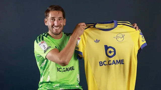 Franco Ravizzoli, sat down while wearing a green Leicester City goalkeeping jersey, holding a yellow Leicester City shirt up while smiling.