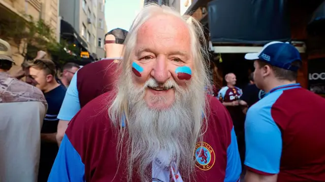 An Aston Villa fan with claret and blue face paint on. The gent has a long white beard and smiles.