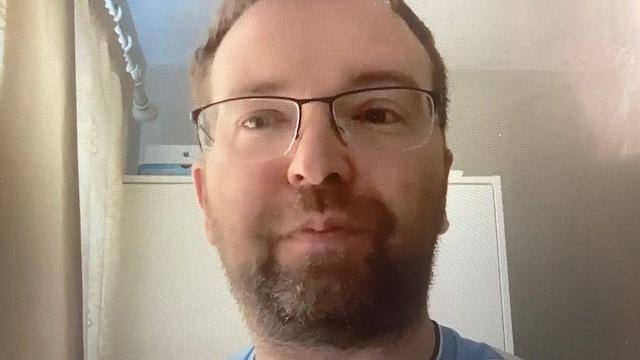A man is wearing a blue top and glasses. He has brown hair and a beard.