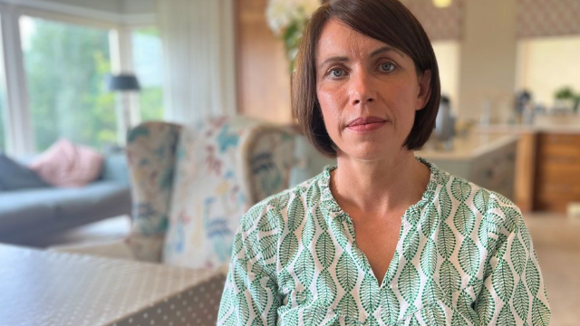 Angela O'Neill, a woman with brown, bobbed hair, looks into the camera.  She is wearing a white top patterned with green leaves.  There is a blurred background of a kitchen-diner behind her, with a large window on the left overlooking trees. 