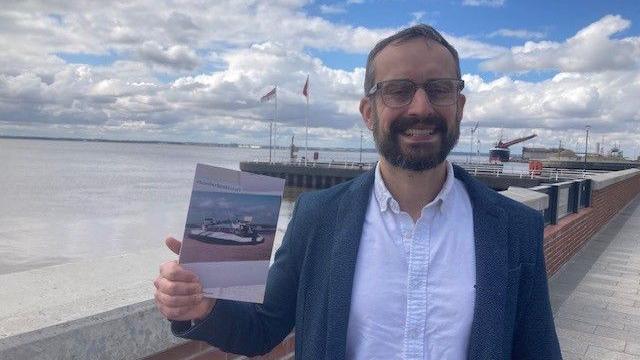 A man with a beard and glasses stands holding a book with a photo of a hovercraft on the front. He is wearing a white shirt and a blue blazer. He is by the harbour wall, with the river Humber in the background. 