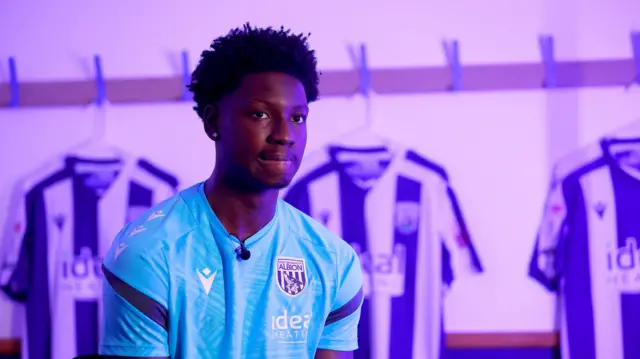 Jamaldeen Jimoh-Aloba being unveiled as the new West Bromwich Albion signing wearing a light blue Albion shirt with home shirts on hangers are lined up in the background