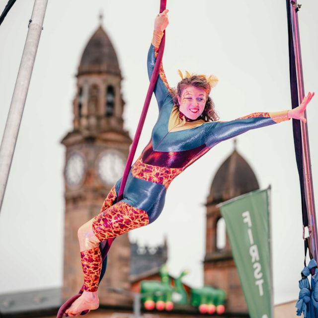 An aerial performer in a blue, orange and pink unitard swings on a rope with Paisley town hall's clock tower in the background.
