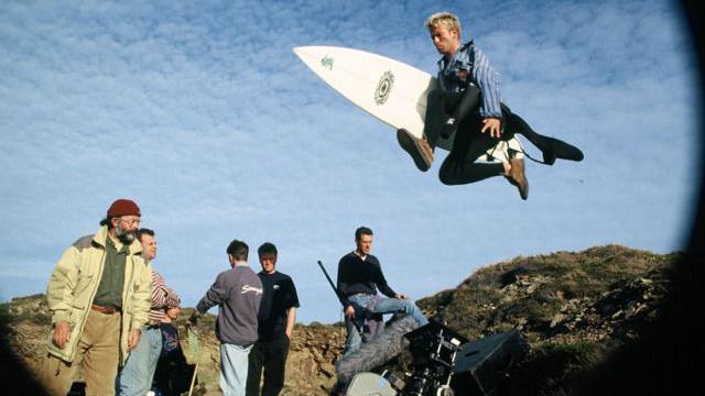 A fish eye camera view of five film cast members and a surfer who is jumping into the air with a surfboard under his arm.