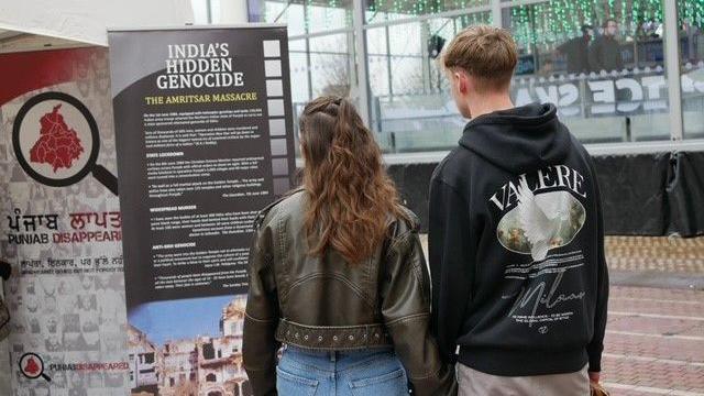 A man and a woman are reading from a black display banner which is titled India's Hidden Genocide. She is wearing a brown leather jacket and blue jeans, he is wearing a black hoodie and beige trousers.