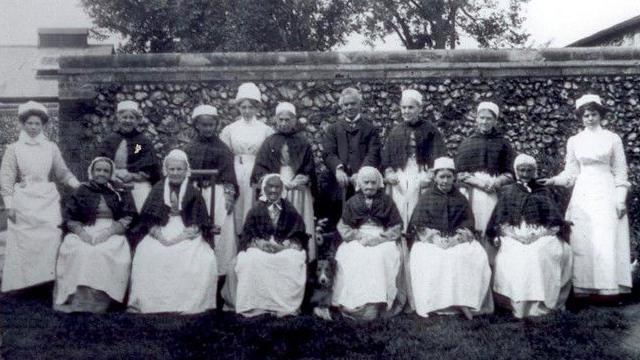 Dr Allan Minns, wearing a dark three piece suit, standing with 11 Thetford Union Workhouse women inmates and three nurses. He is in the back row, standing, wearing a dark three piece suit. The women inmates are wearing shawls over light-coloured dresses and light coloured caps on their hair. There are also three nurses, two at either end and one standing towards the middle