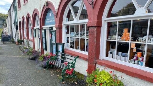 The new tourist information centre. The building has large, arched windows running along the length of the building, which are framed with red-painted stonework. Mugs and souvenirs are displayed in the window. A green bench and flowers are displayed outside.