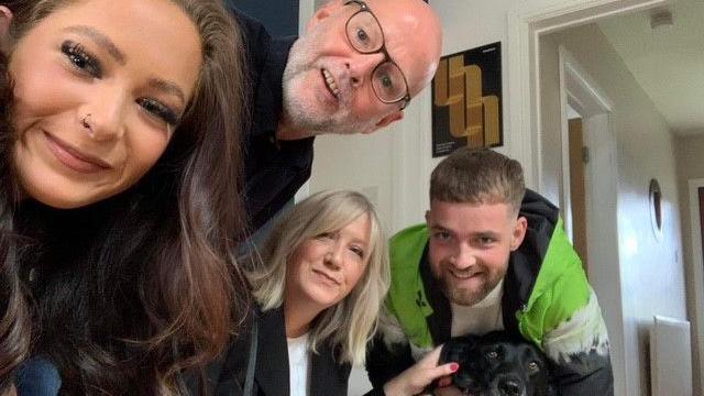 A group of people are gathered indoors near a doorway, leaning toward the camera. They are dressed casually, and one person is wearing a bright green and black jacket. In the center, a black dog with a shiny coat is sitting on the floor, looking toward the camera.